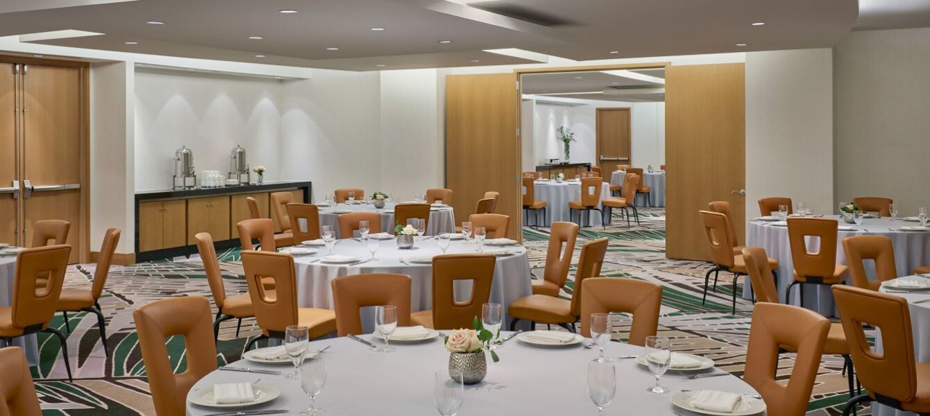 A banquet room with round tables set for dining, tan chairs, white tablecloths, glassware, and wooden doors in a modern, well-lit space.
