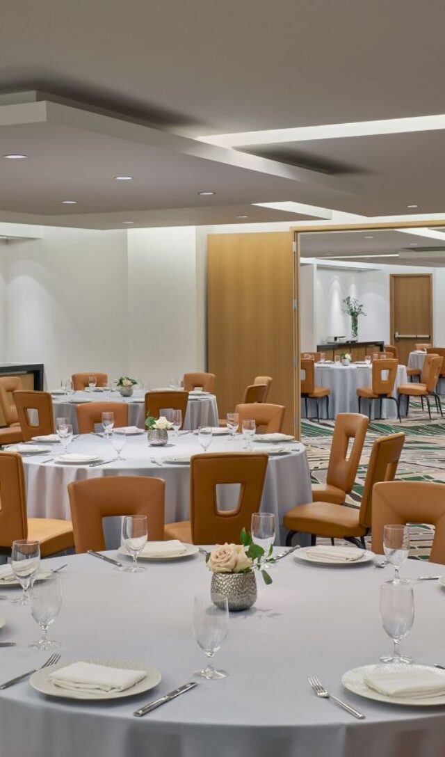 A banquet room with round tables set for dining, tan chairs, white tablecloths, glassware, and wooden doors in a modern, well-lit space.