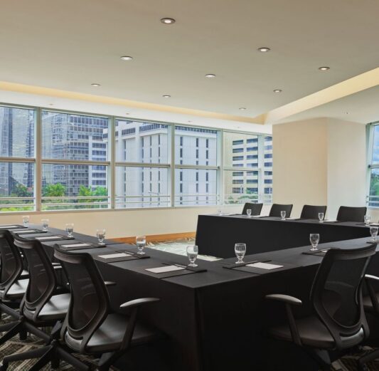 Modern conference room with U-shaped table setup, black chairs, notepads, and glasses, large windows showing a cityscape with tall buildings.