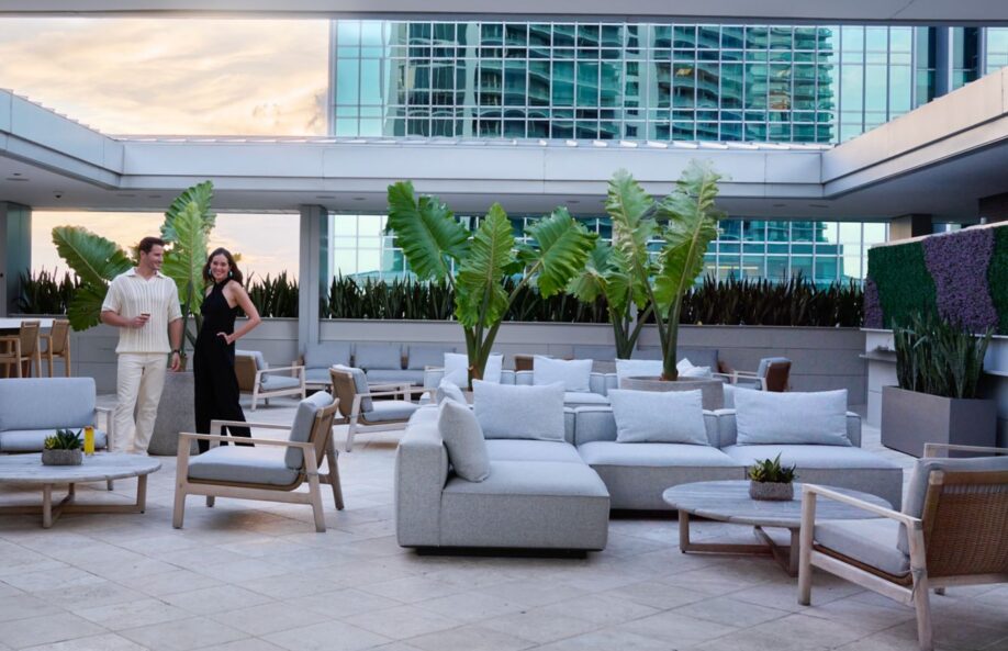 A man and woman stand and smile on a modern outdoor terrace with gray lounge furniture, large green plants, and tall glass buildings in the background.