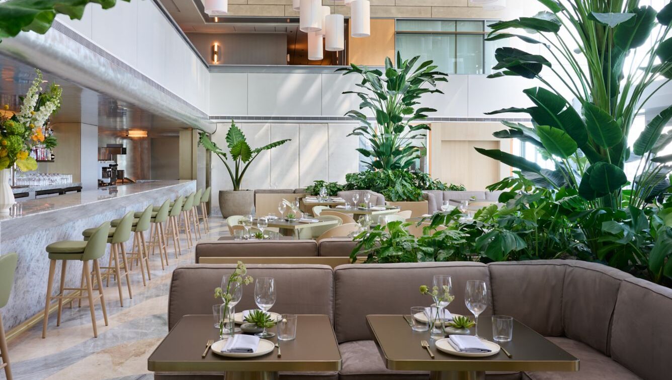 Modern restaurant with green plants, elegant tables set for dining, a marble bar with high chairs, and natural light from large windows.