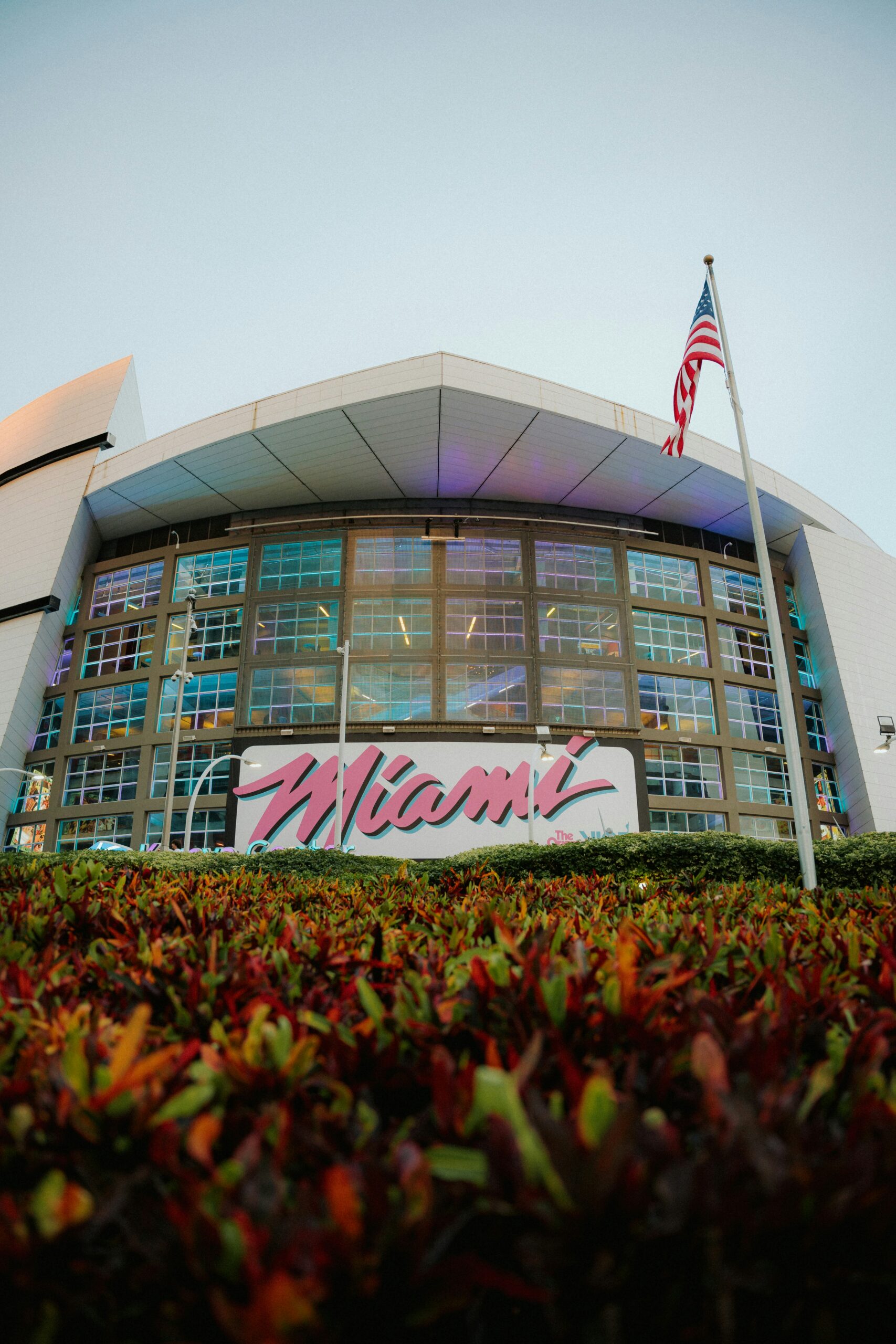 Front view of an arena with a large “Miami” sign, glass windows, an American flag, and bushes in the foreground under a clear sky.