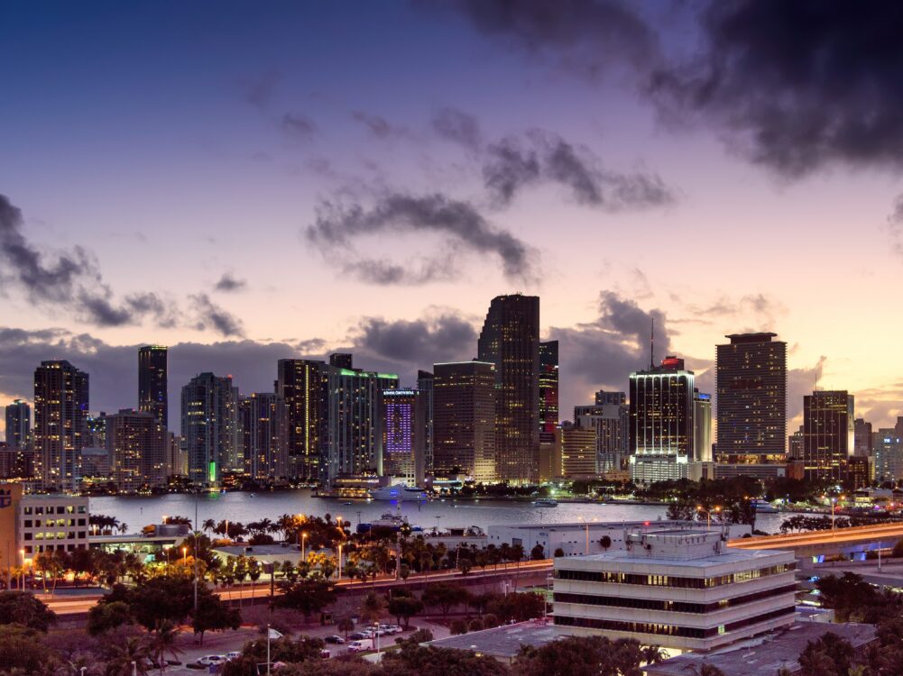 Miami skyline at dusk with illuminated buildings, palm trees, and cloudy sky reflected on the bay.