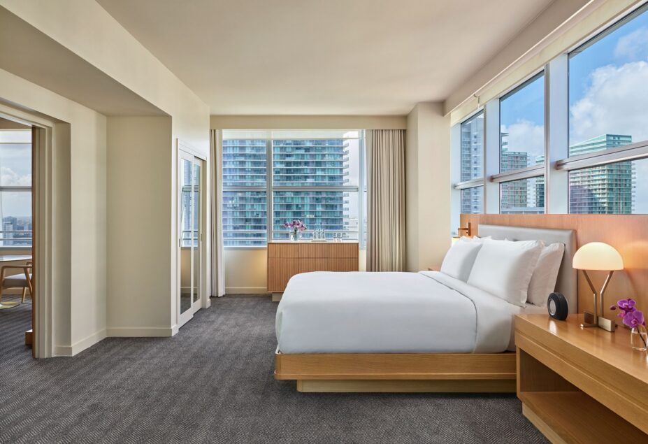 Modern hotel room with a large bed, bedside tables, big windows with city views, gray carpet, light walls, and a desk area in an adjacent room.