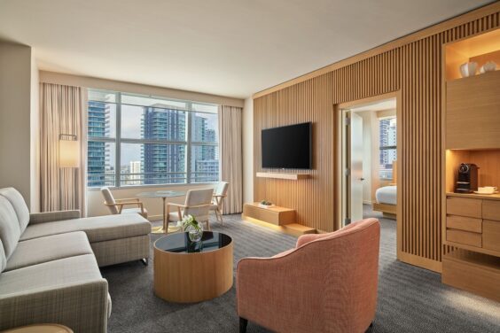 Modern hotel suite with a sofa, armchair, TV, round table, large window with city views, and a visible bedroom through an open doorway.