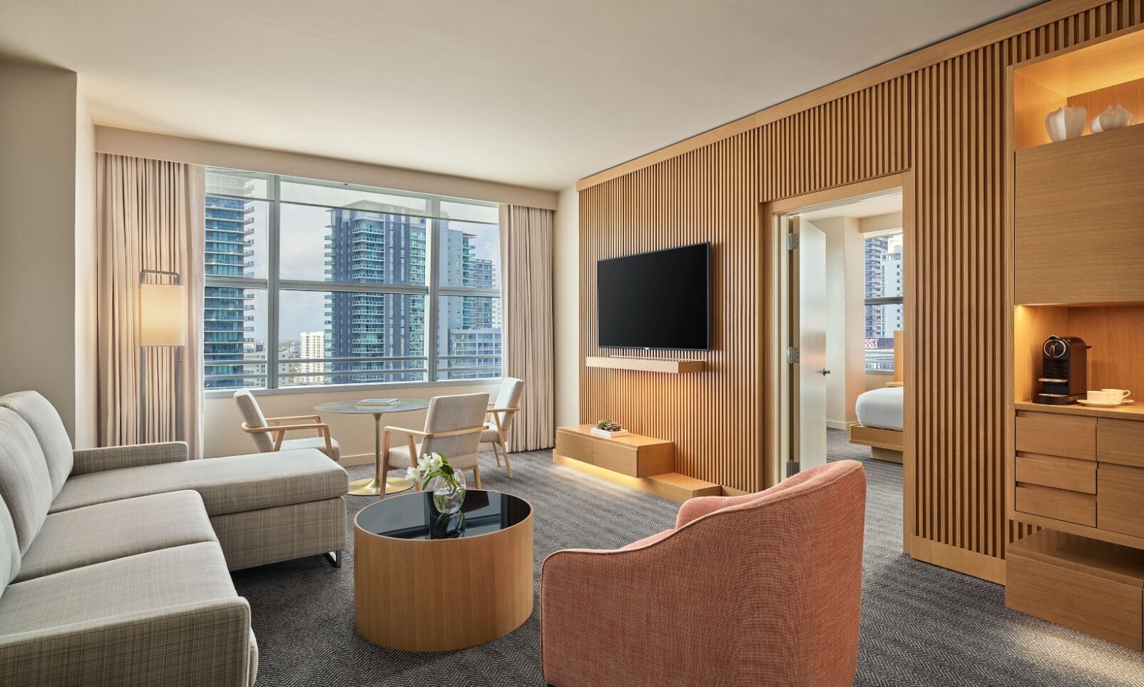 Modern hotel suite with a sofa, armchair, TV, desk, and large window overlooking city skyscrapers; adjoining bedroom partially visible.