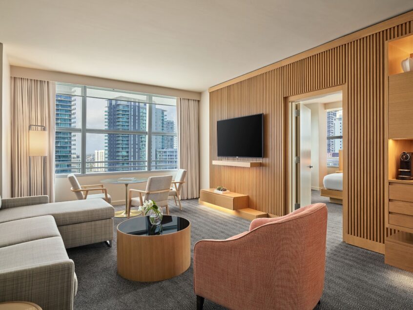 Modern hotel suite with a sofa, armchair, TV, desk, and large window overlooking city skyscrapers; adjoining bedroom partially visible.