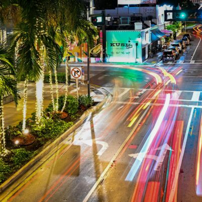 A city street at night with palm trees wrapped in lights and colorful car light trails curving around a corner, creating a lively urban scene.